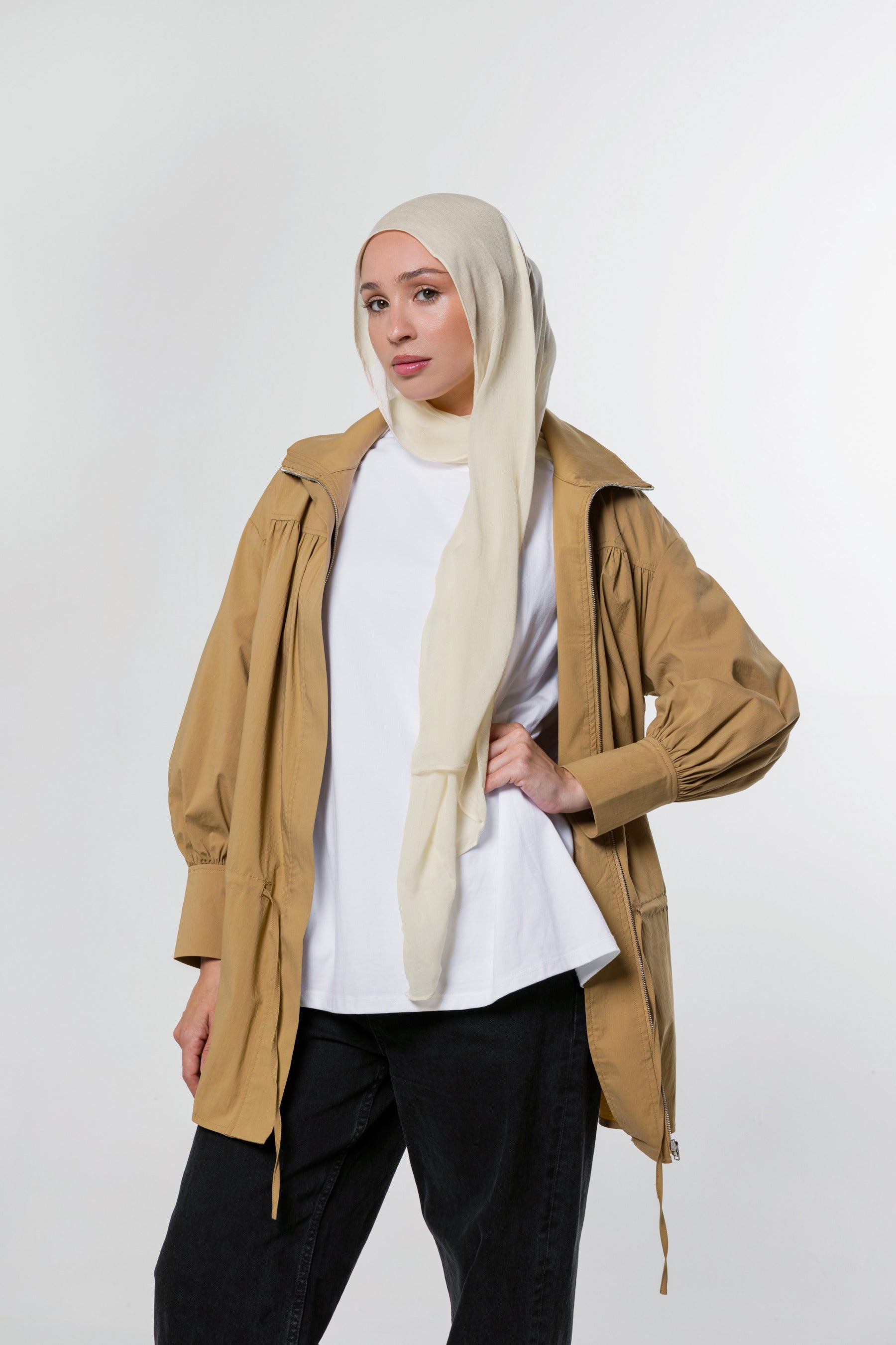 A woman modeling a tan hooded jacket over a white top and black pants, wearing a light beige hijab, against a plain light gray studio background.
