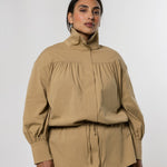 A model wearing a beige high-neck, long-sleeve blouse with gathered detailing and a drawstring waist, paired with a dark gray skirt, against a light gray studio background.