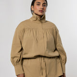 A model wearing a beige high-neck, long-sleeve blouse with gathered detailing and a drawstring waist, paired with a dark gray skirt, against a light gray studio background.