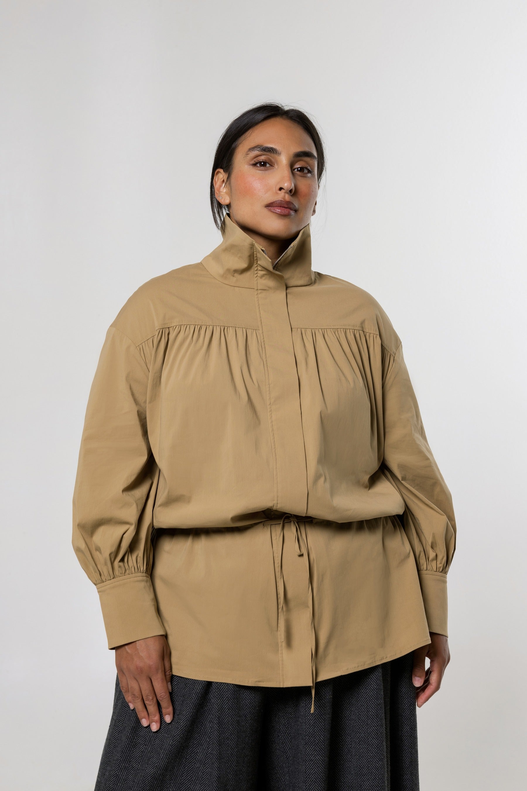 A model wearing a beige high-neck, long-sleeve blouse with gathered detailing and a drawstring waist, paired with a dark gray skirt, against a light gray studio background.