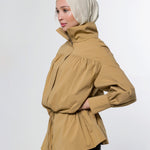 A woman wearing a beige hijab and a tan high-neck, long-sleeved wrap jacket with a gathered waist, shown in profile against a light gray studio background.