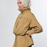 A woman wearing a beige hijab and a tan high-neck, long-sleeved wrap jacket with a gathered waist, shown in profile against a light gray studio background.
