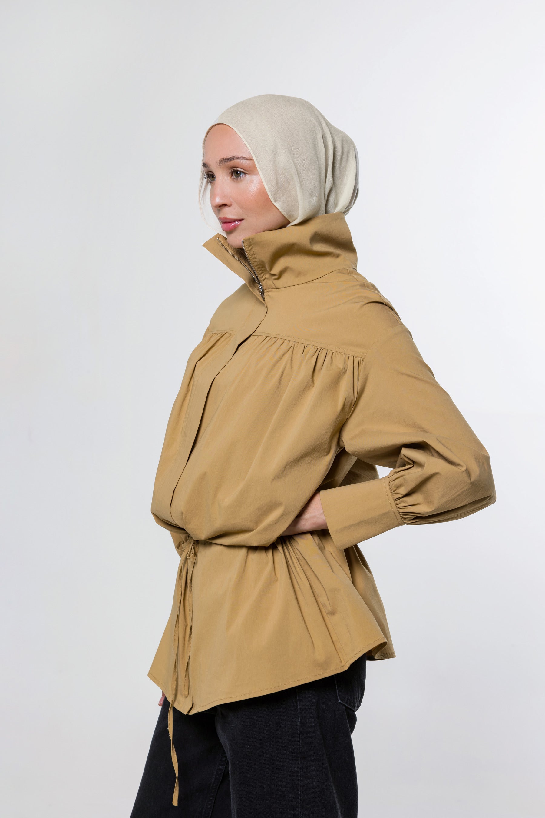 A woman wearing a beige hijab and a tan high-neck, long-sleeved wrap jacket with a gathered waist, shown in profile against a light gray studio background.