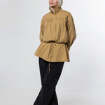 A woman wearing a tan long-sleeve peplum blouse with a drawstring waist, black wide-leg pants, and a light beige hijab, standing against a plain light gray studio background.