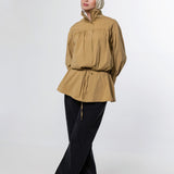 A woman wearing a tan long-sleeve peplum blouse with a drawstring waist, black wide-leg pants, and a light beige hijab, standing against a plain light gray studio background.