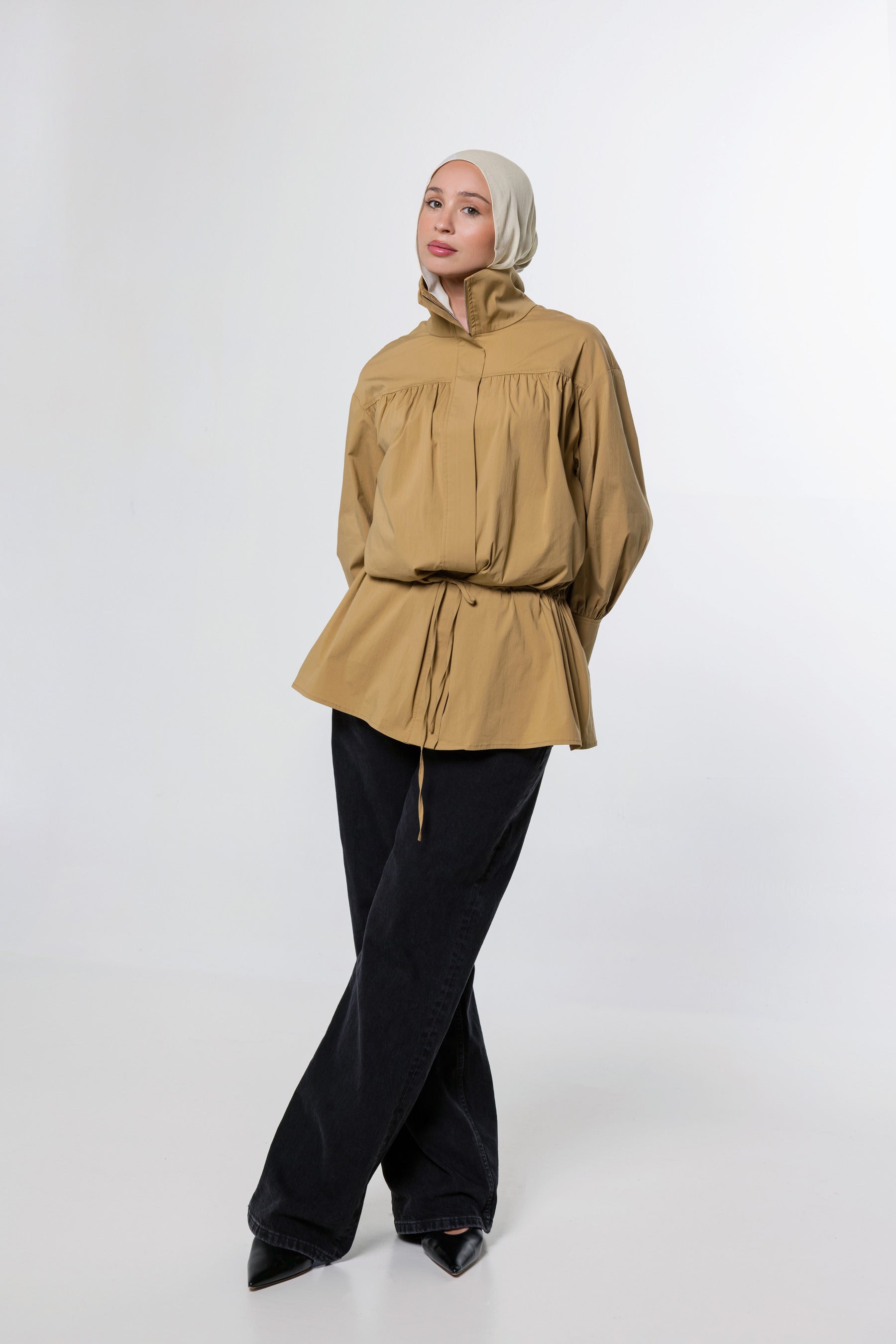 A woman wearing a tan long-sleeve peplum blouse with a drawstring waist, black wide-leg pants, and a light beige hijab, standing against a plain light gray studio background.
