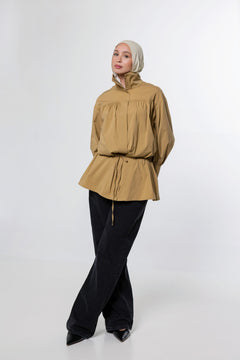 A woman wearing a tan long-sleeve peplum blouse with a drawstring waist, black wide-leg pants, and a light beige hijab, standing against a plain light gray studio background.