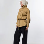 A model wearing a tan high-neck, long-sleeve hooded jacket with a drawstring waist, paired with black wide-leg trousers and a light beige hijab, standing against a white studio background.