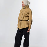 A model wearing a tan high-neck, long-sleeve hooded jacket with a drawstring waist, paired with black wide-leg trousers and a light beige hijab, standing against a white studio background.