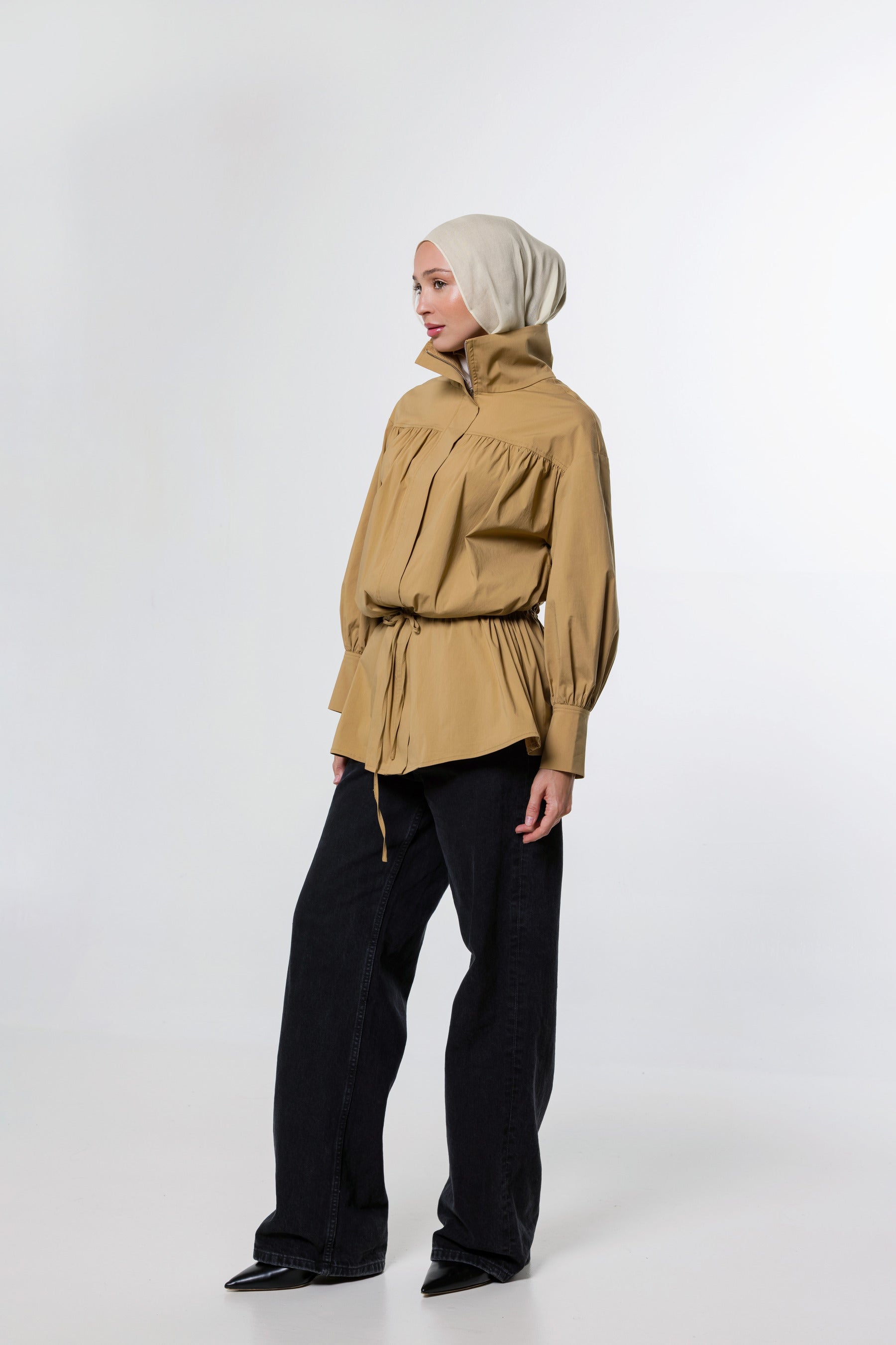 A model wearing a tan high-neck, long-sleeve hooded jacket with a drawstring waist, paired with black wide-leg trousers and a light beige hijab, standing against a white studio background.