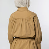 A person shown from the back wearing a tan long-sleeve blouse with a gathered waist and a light beige hijab, photographed against a plain light gray studio background.