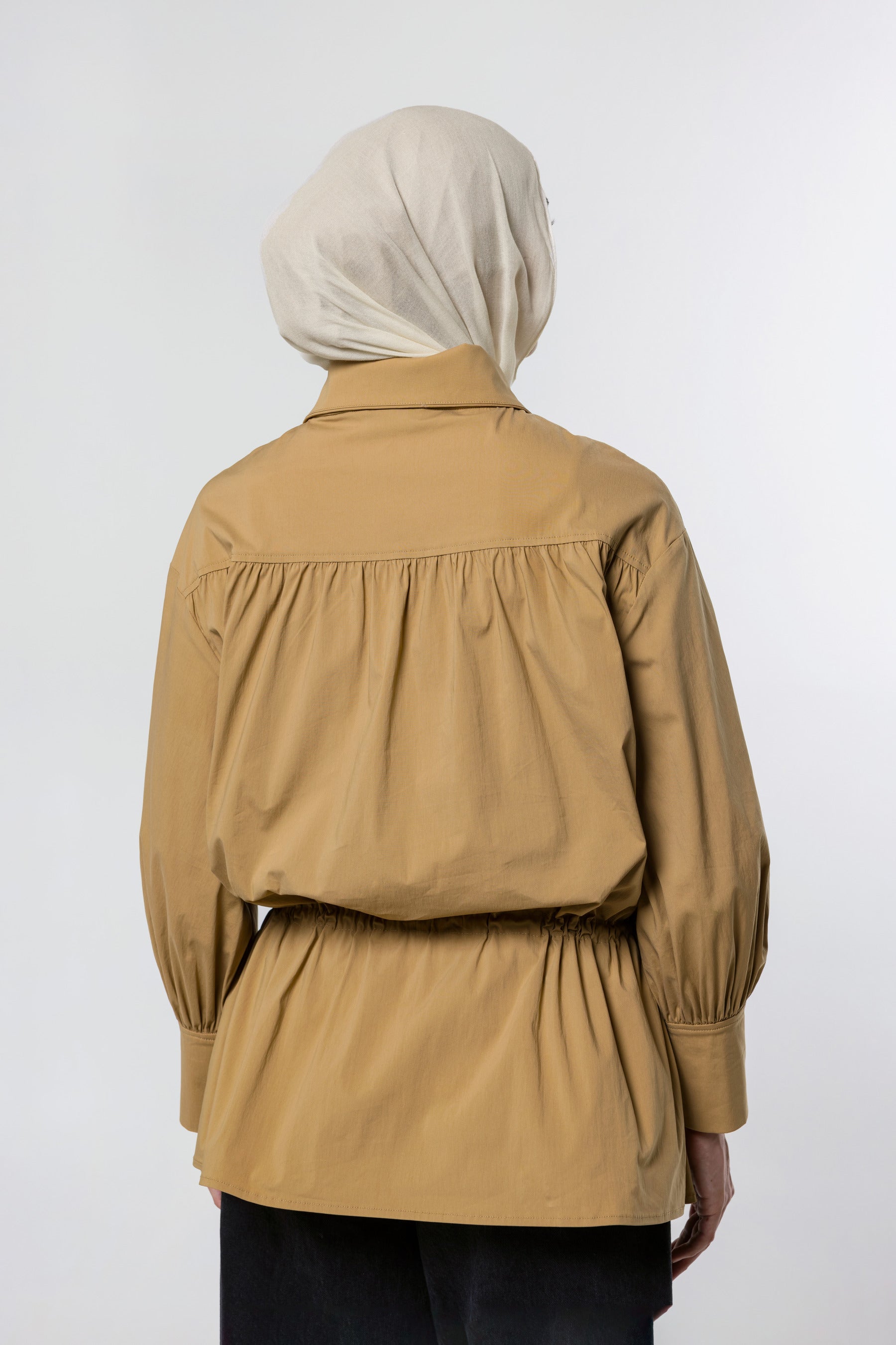 A person shown from the back wearing a tan long-sleeve blouse with a gathered waist and a light beige hijab, photographed against a plain light gray studio background.