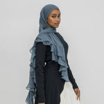 A woman wearing a slate blue ruffled hijab draped over her shoulders, paired with a black long-sleeve dress and a white skirt, standing against a light gray studio background.