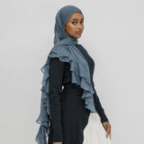 A woman wearing a slate blue ruffled hijab draped over her shoulders, paired with a black long-sleeve dress and a white skirt, standing against a light gray studio background.