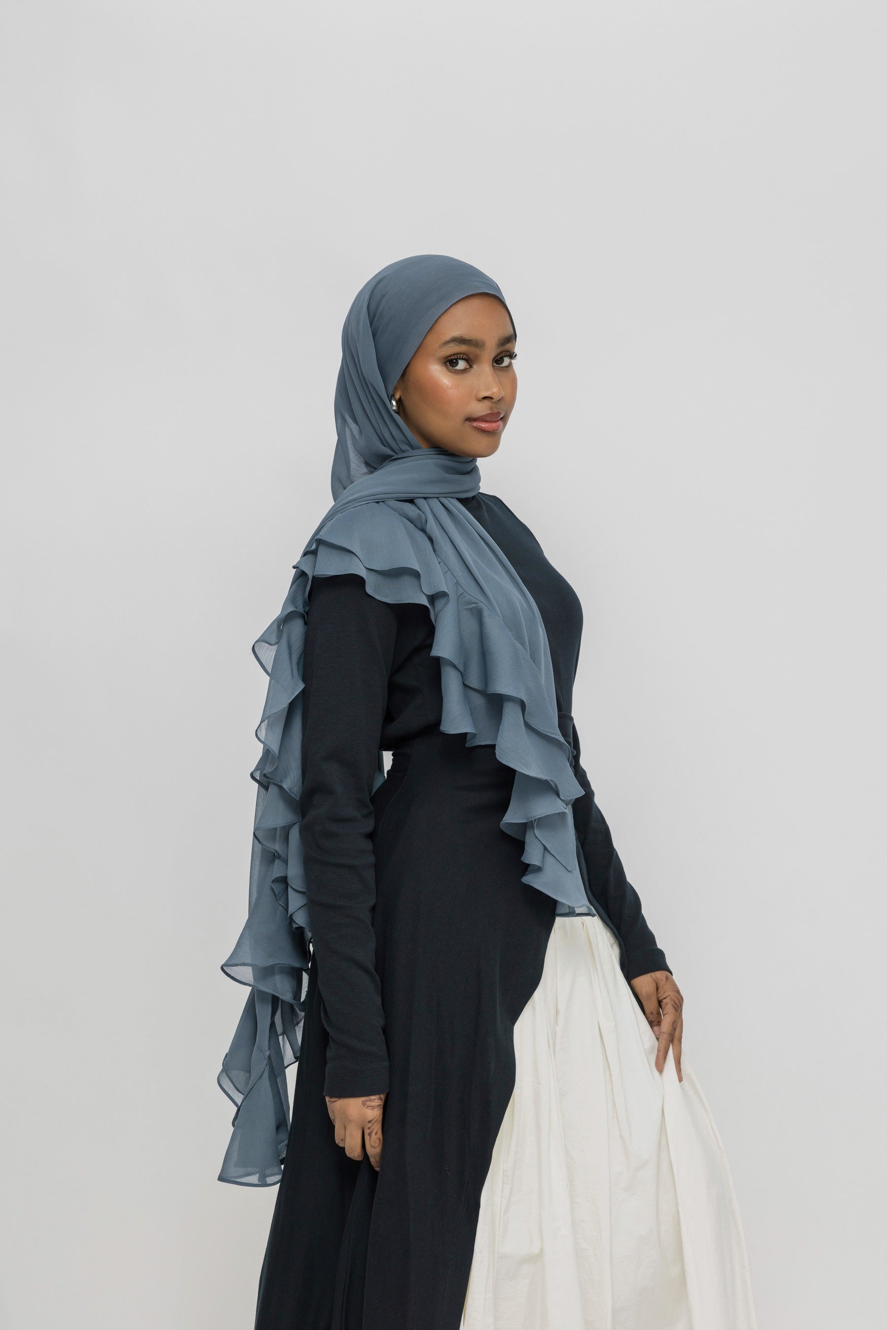 A woman wearing a slate blue ruffled hijab draped over her shoulders, paired with a black long-sleeve dress and a white skirt, standing against a light gray studio background.