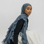 A woman wearing a slate blue ruffled hijab and a black long-sleeve top, seated on a white draped fabric against a light gray studio background.