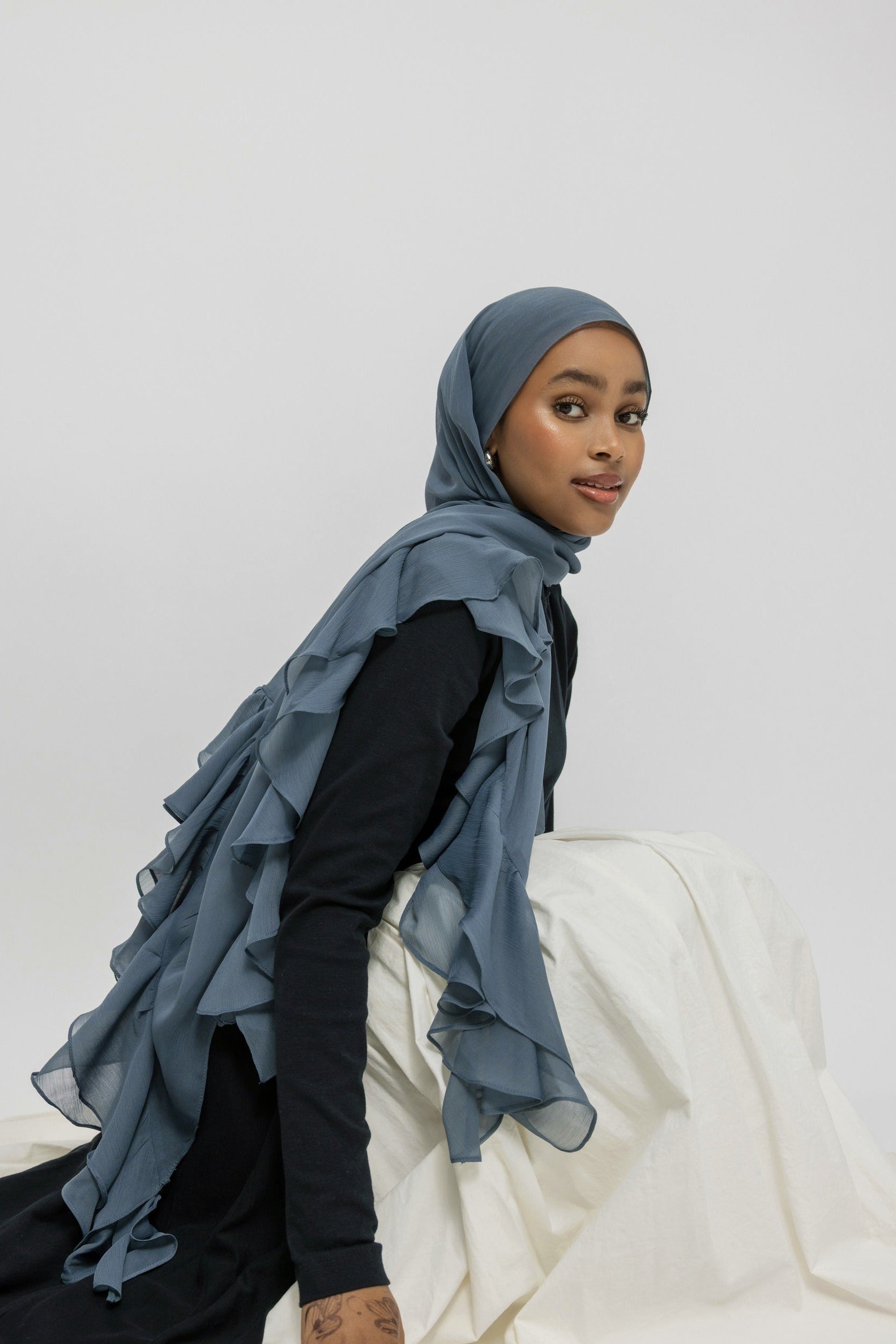 A woman wearing a slate blue ruffled hijab and a black long-sleeve top, seated on a white draped fabric against a light gray studio background.