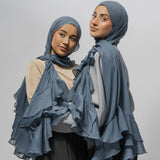 Two women wearing matching ruffled hijabs and draped shawls in a dusty slate blue color, posed against a solid light gray studio background.