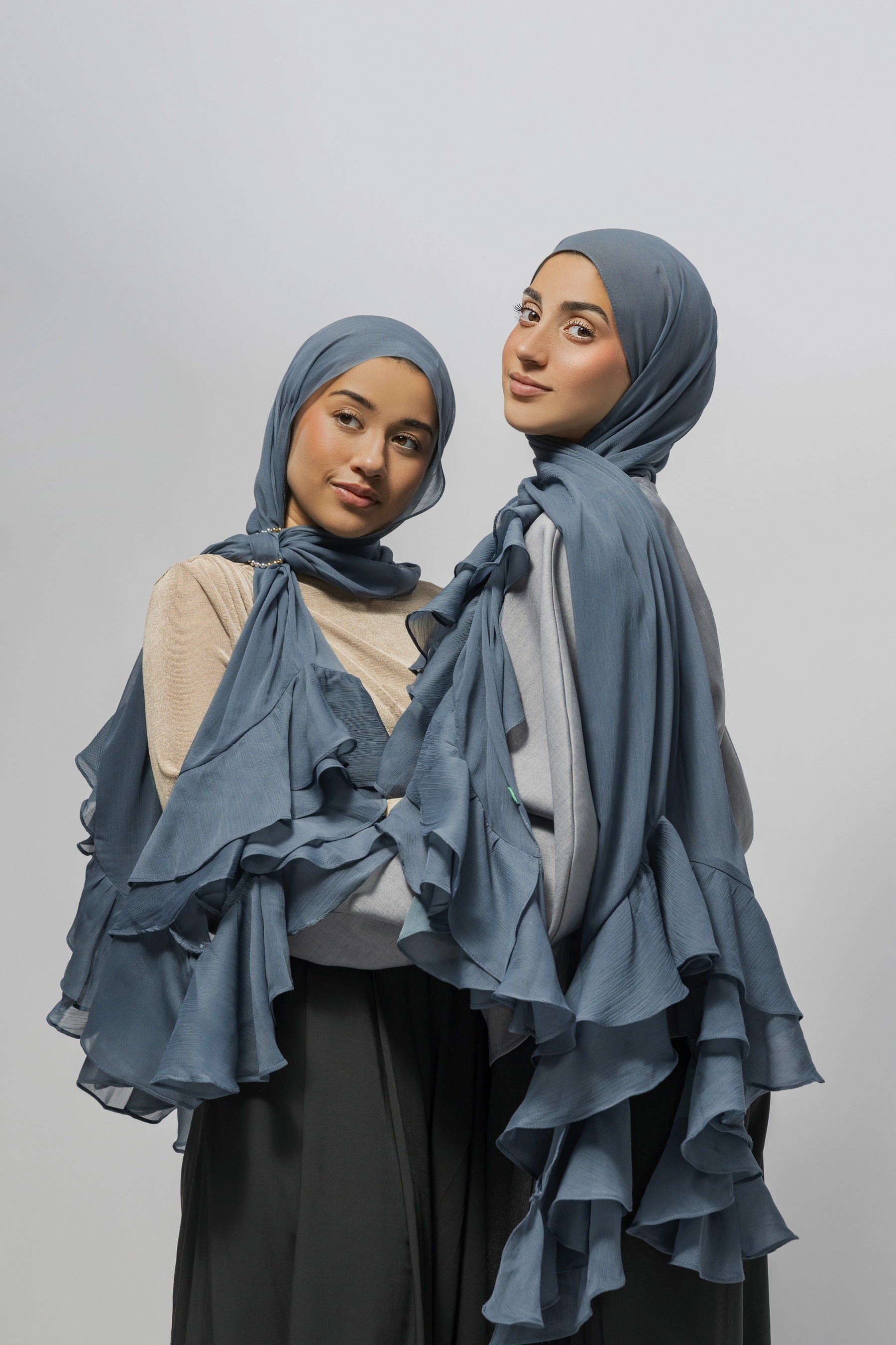 Two women wearing matching ruffled hijabs and draped shawls in a dusty slate blue color, posed against a solid light gray studio background.