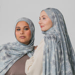 Two women wearing matching light blue hijabs with floral patterns against a plain background