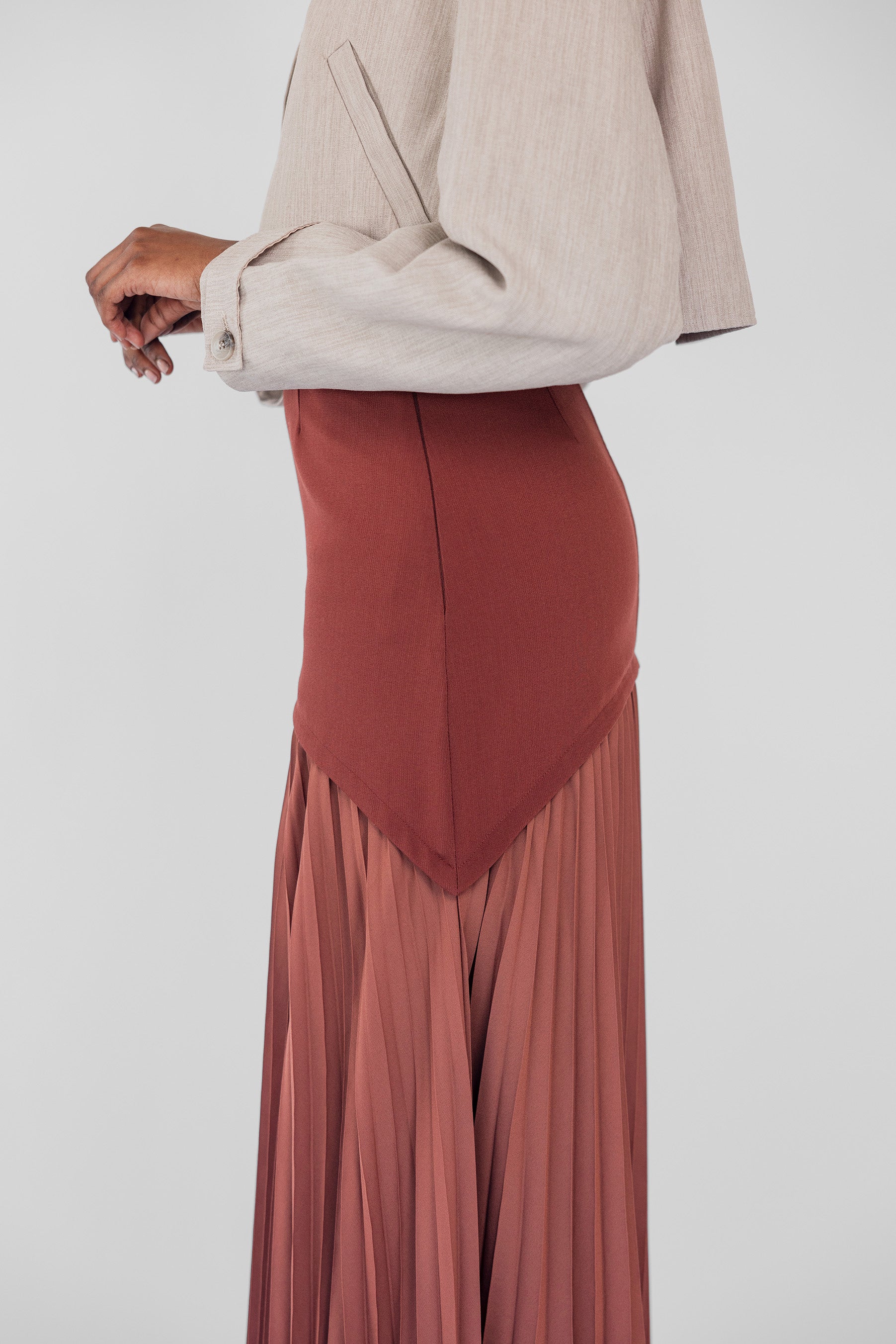 Person wearing a beige blazer and rust-colored pleated skirt on a white background