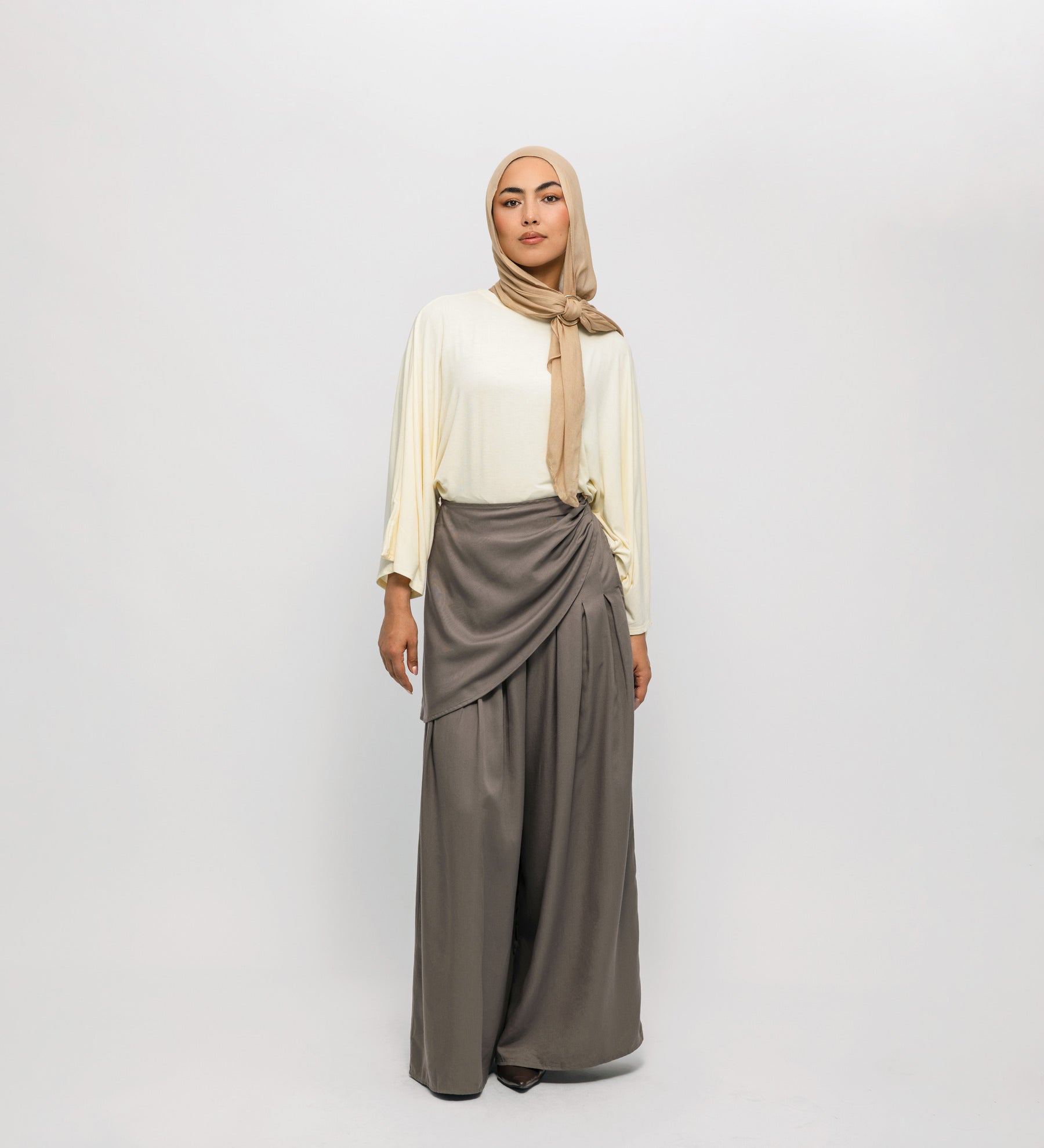 Woman wearing a beige hijab, white top, and gray skirt on a white background