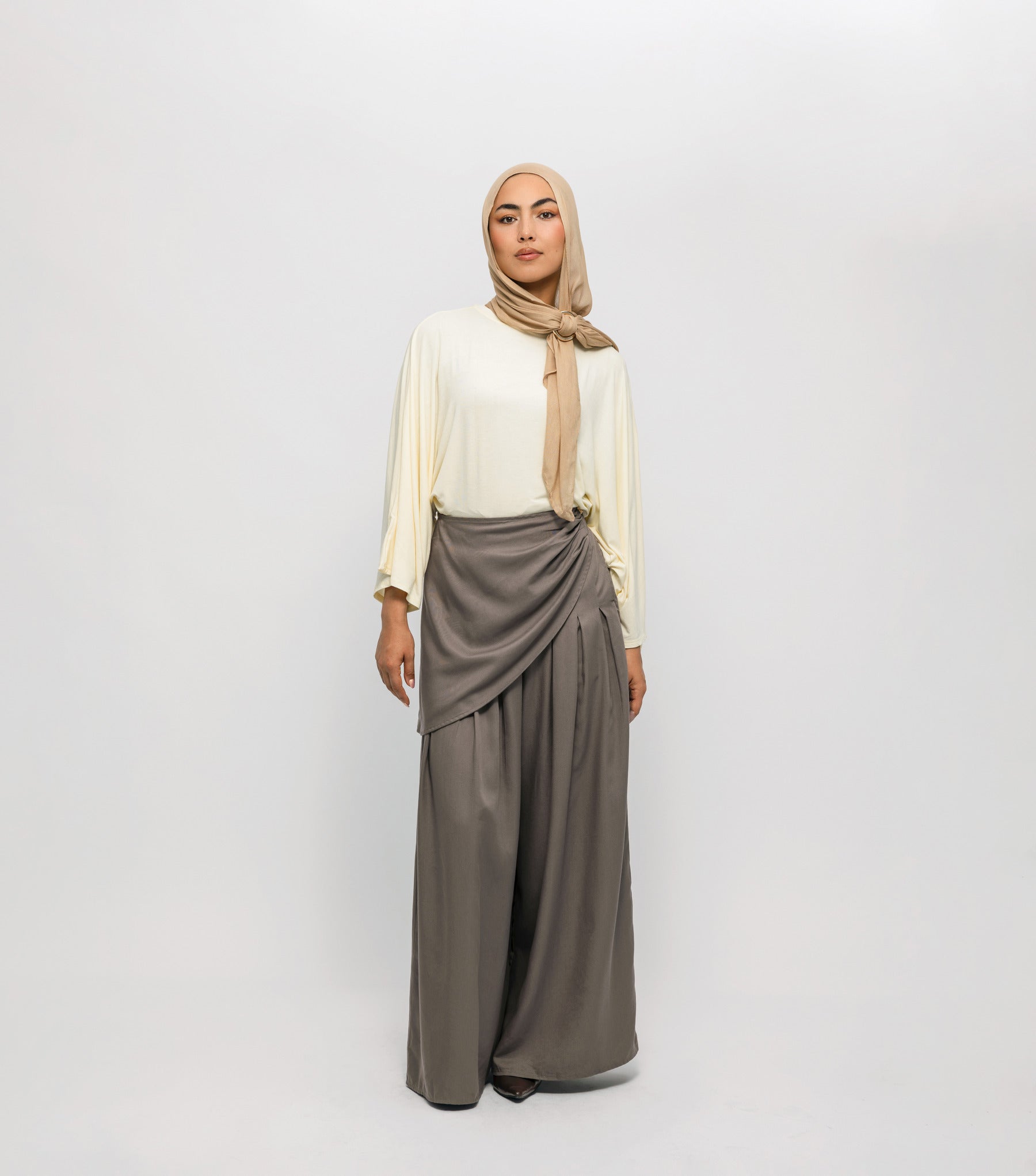 Woman wearing a beige hijab, white top, and gray skirt on a white background