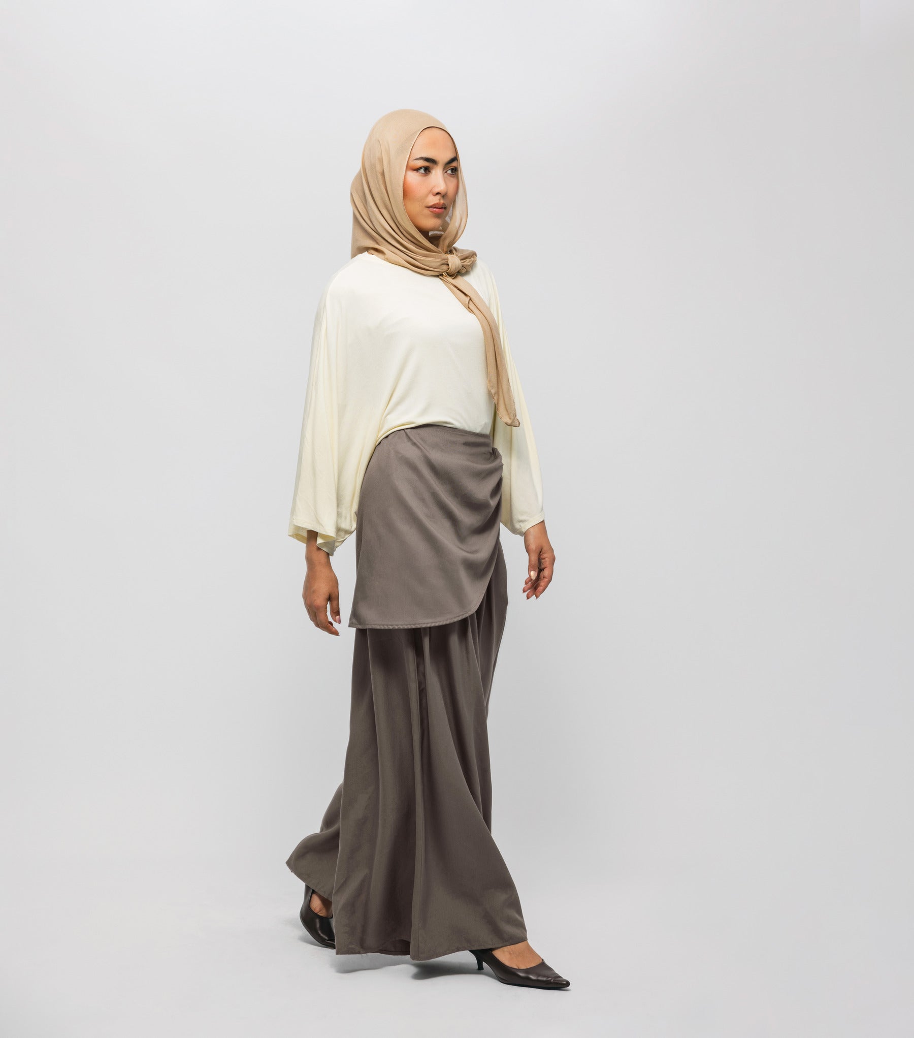 Woman wearing a beige hijab, cream top, and brown skirt on a white background