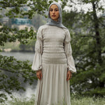 Woman in a light-colored dress and hijab standing in a natural setting with trees and water.