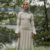 Woman in a light-colored dress and hijab standing in a natural setting with trees and water.