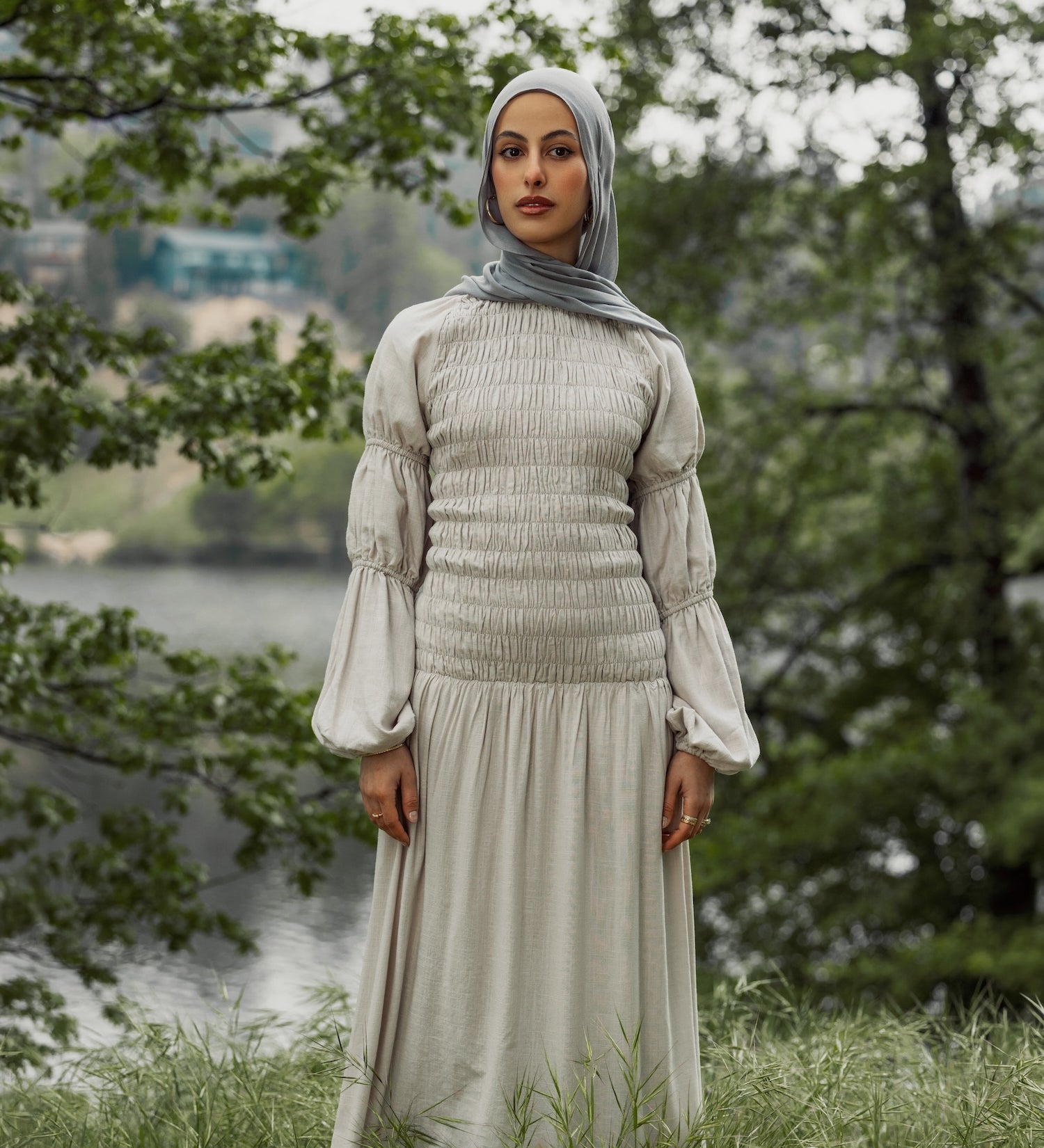 Woman in a light-colored dress and hijab standing in a natural setting with trees and water.