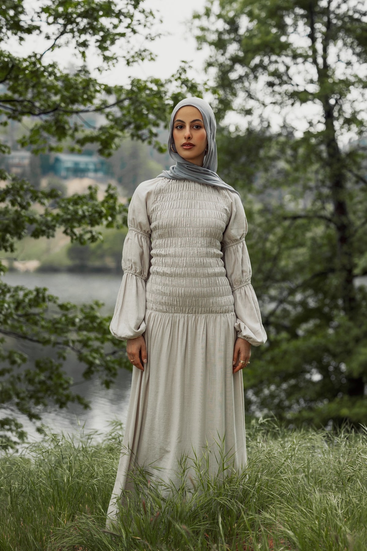 Woman in a light-colored dress and hijab standing in a natural setting with trees and water.