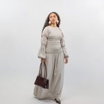 Woman wearing a light gray smocked dress holding a brown handbag on a white background