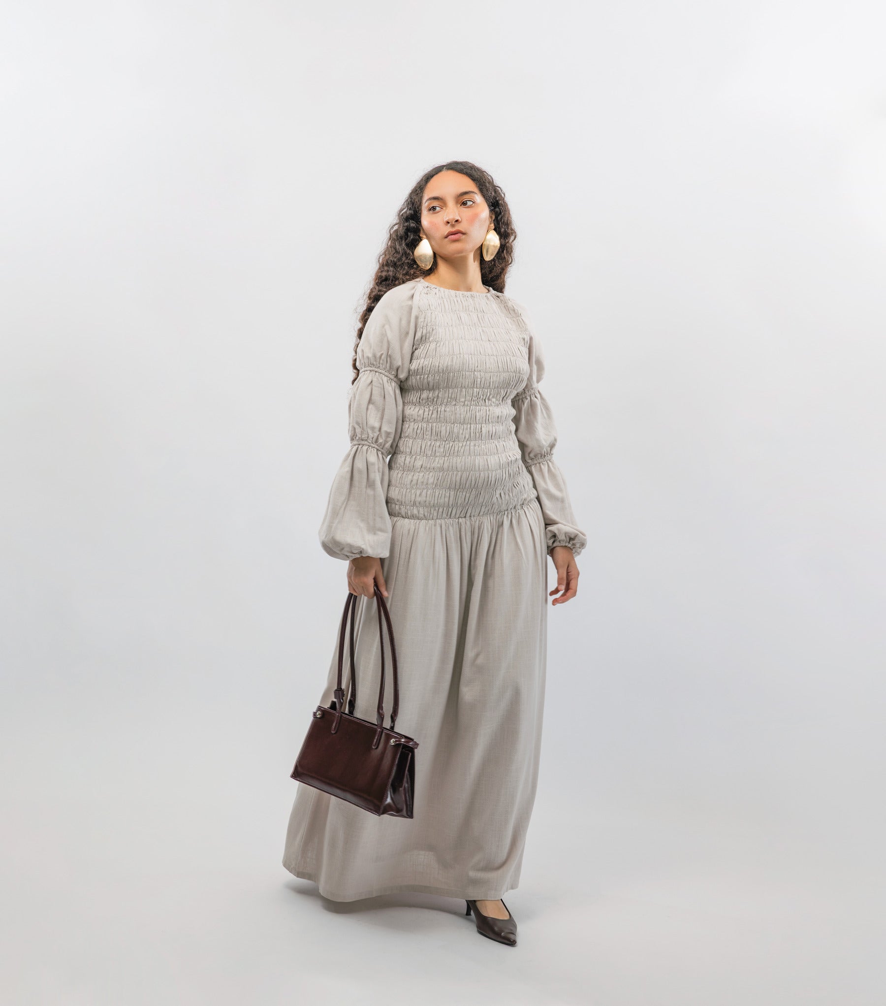 Woman wearing a light gray smocked dress holding a brown handbag on a white background