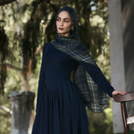 A woman wearing a dark navy long-sleeve maxi dress and matching hijab with a plaid pattern, standing in an outdoor garden setting next to a wooden chair.