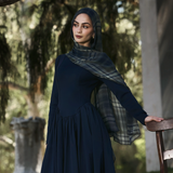 A woman wearing a dark navy long-sleeve maxi dress and matching hijab with a plaid pattern, standing in an outdoor garden setting next to a wooden chair.
