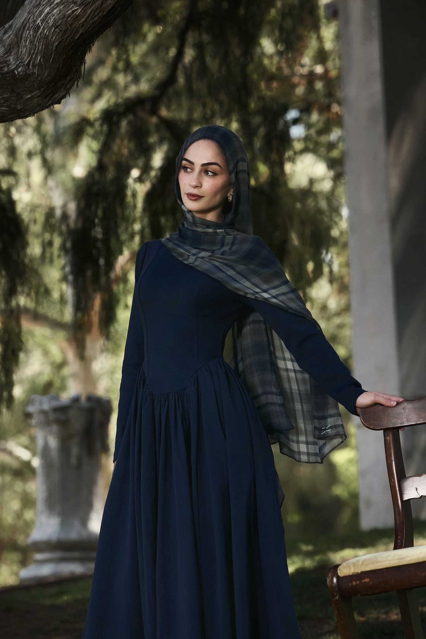 A woman wearing a dark navy long-sleeve maxi dress and matching hijab with a plaid pattern, standing in an outdoor garden setting next to a wooden chair.