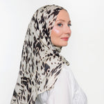 A woman wearing a lightweight patterned hijab in beige, black, and white, paired with a white long-sleeve top, shown in profile against a plain white background.