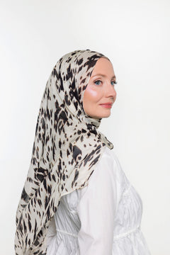 A woman wearing a lightweight patterned hijab in beige, black, and white, paired with a white long-sleeve top, shown in profile against a plain white background.