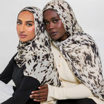 Two women wearing matching beige and black animal-print hijabs, posed together against a plain light gray studio background.