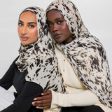 Two women wearing matching beige and black animal-print hijabs, posed together against a plain light gray studio background.