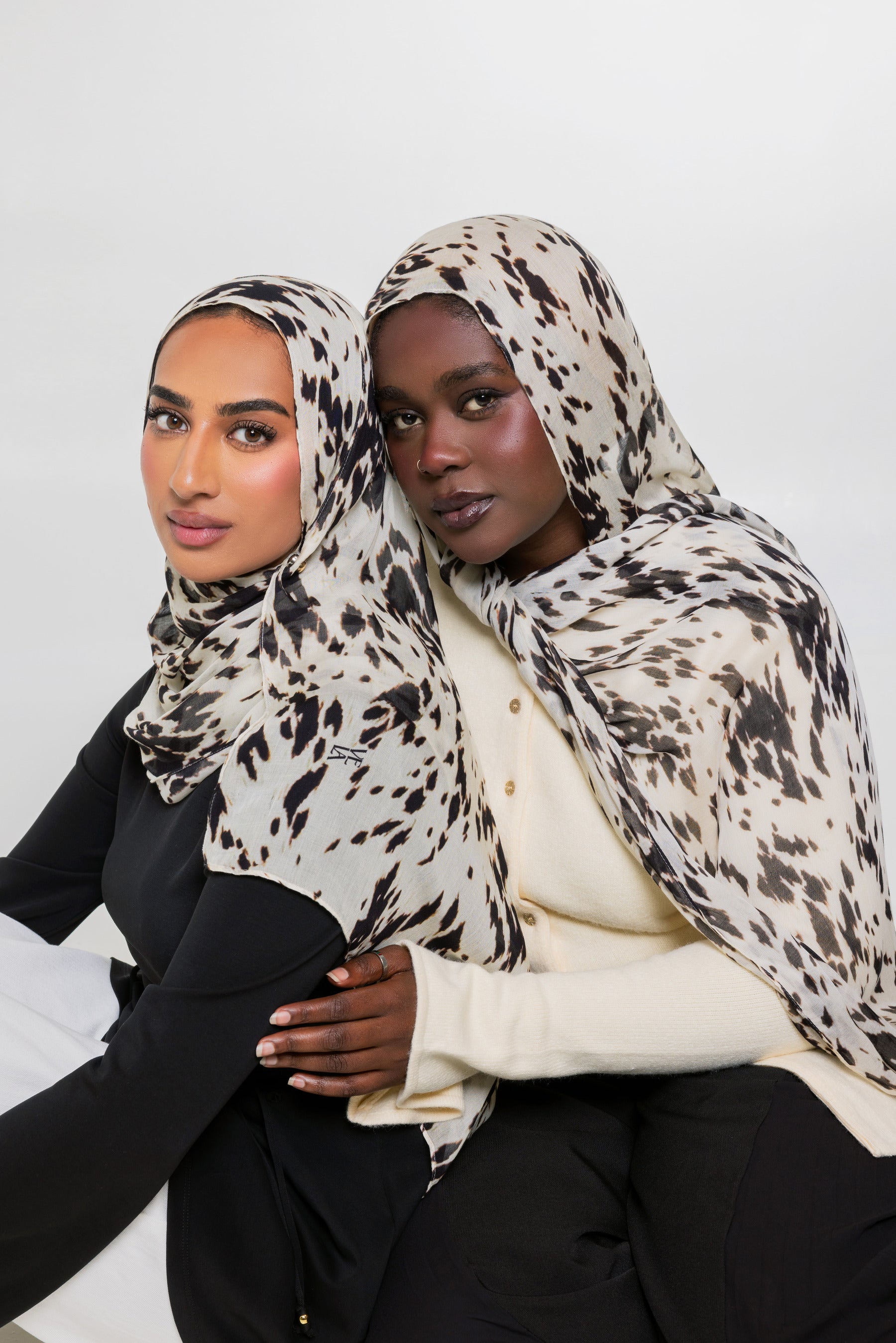 Two women wearing matching beige and black animal-print hijabs, posed together against a plain light gray studio background.
