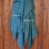Color comparison of two teal toned hijabs labeled 'Storm Blue' and 'Meadow Teal' on a wooden background.