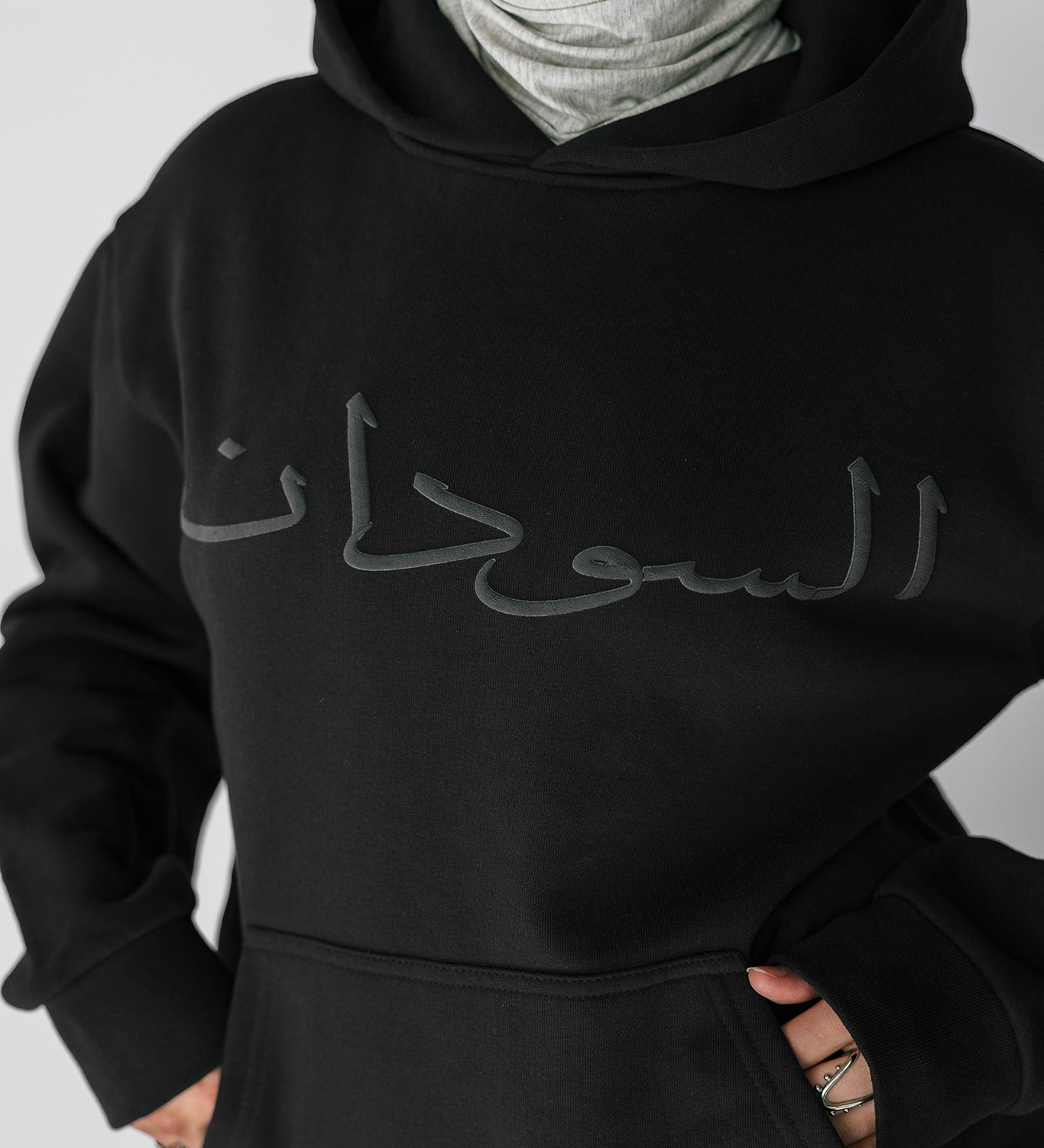 Person wearing a black hoodie with Arabic text on a plain background