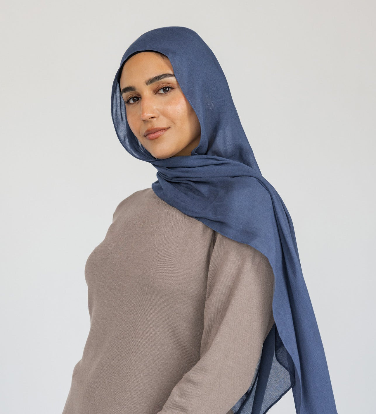 Woman styled in a blue hijab, beige top, and black pants on a white background.