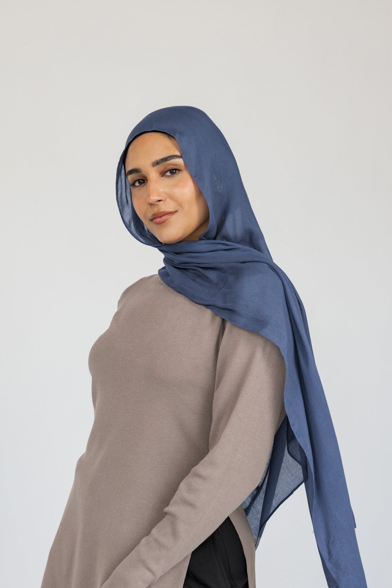 Woman styled in a blue hijab, beige top, and black pants on a white background.