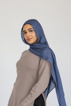 Woman styled in a blue hijab, beige top, and black pants on a white background.