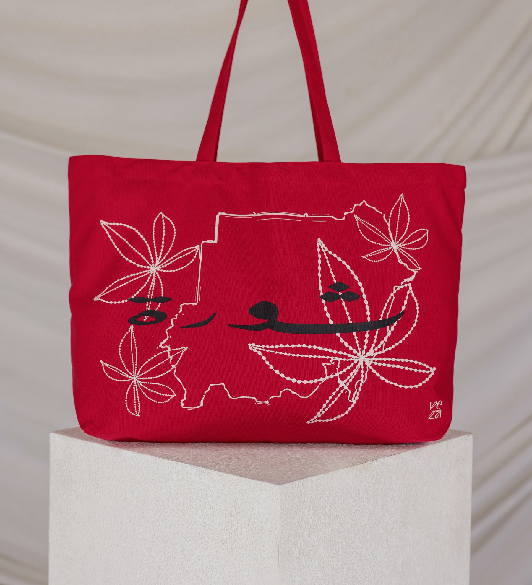 Red tote bag with a map embroidered design on a white pedestal against a beige background