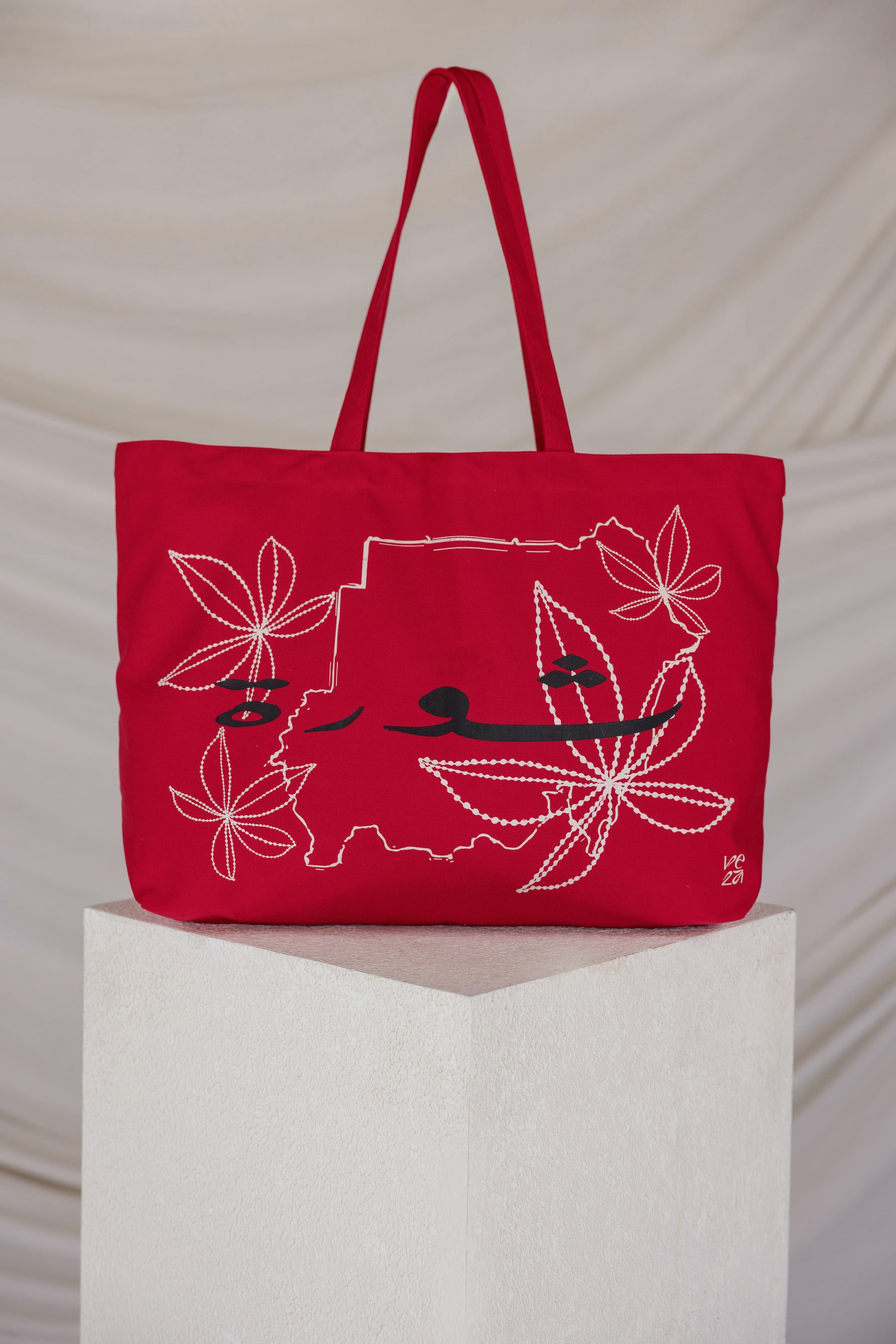 Red tote bag with a map embroidered design on a white pedestal against a beige background