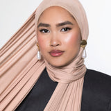 A woman wearing a pleated, draped hijab in a dusty blush pink color, paired with a black top and gold and pearl earrings, set against a plain light gray background.